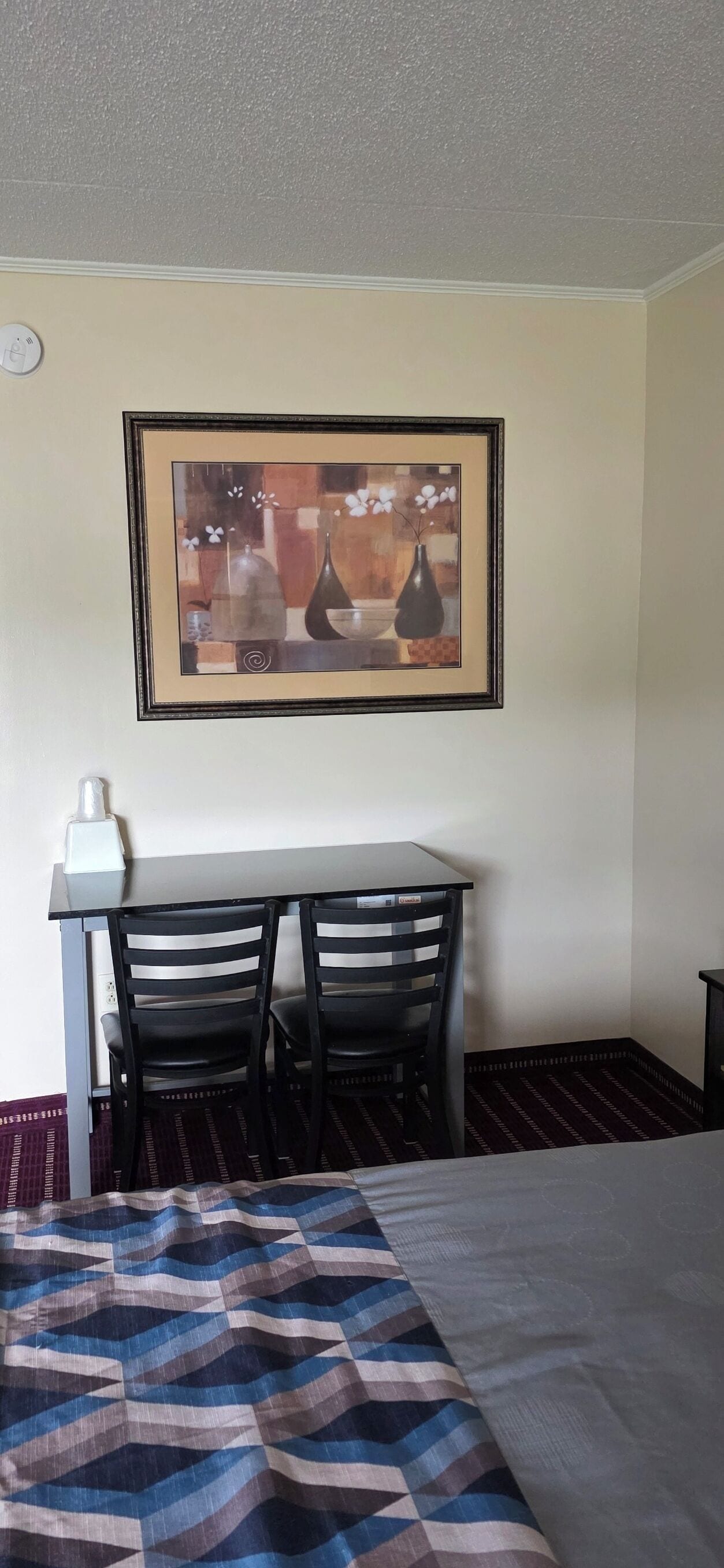 Una camera d'albergo con un letto, una scrivania con due sedie, un quadro appeso al muro e un comodino con una lampada.