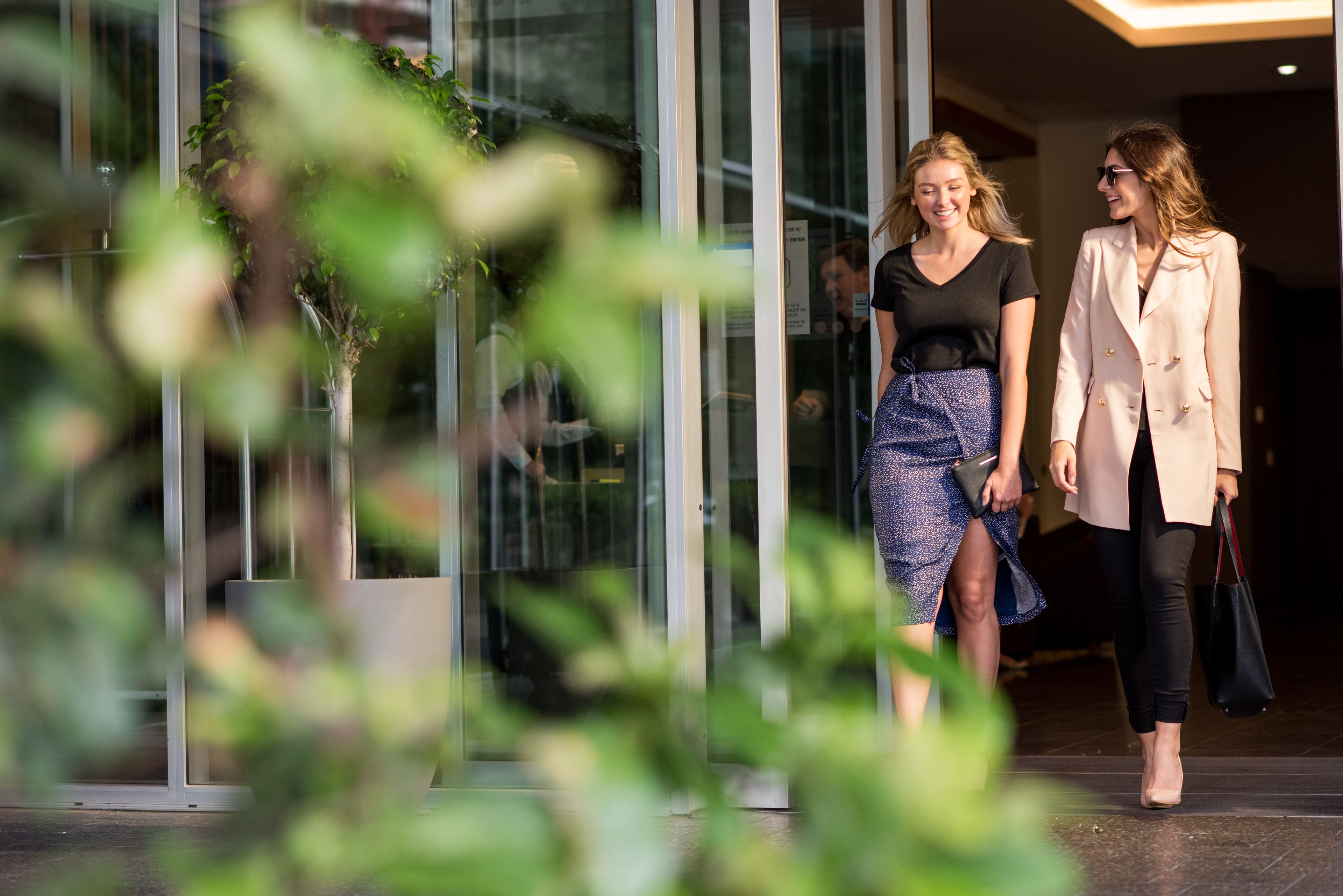 Two individuals walking towards a building entrance with glass doors and a potted plant in the foreground.