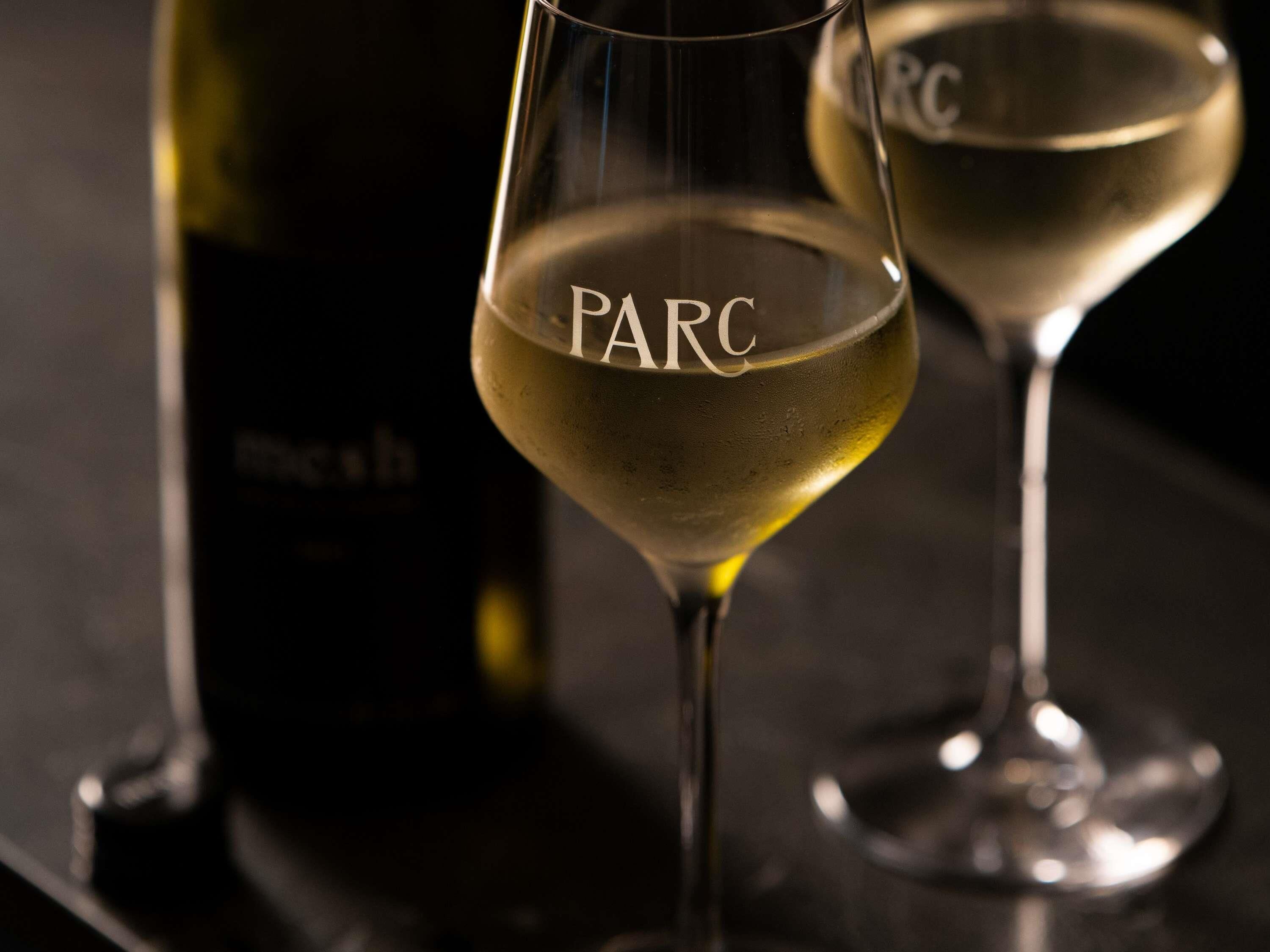 Deux verres de vin blanc, dont l’étiquette porte l’inscription « PARC », posés sur une surface sombre.