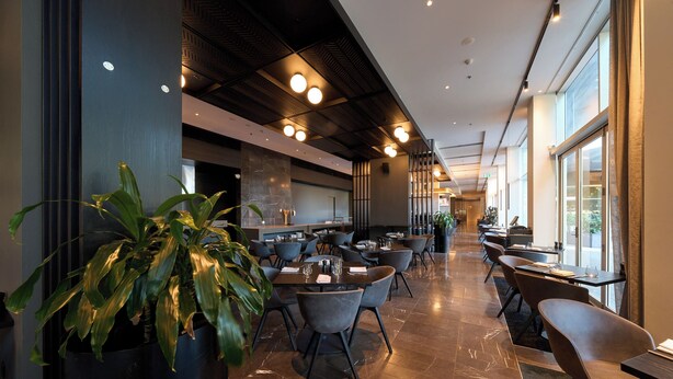 A modern restaurant with a long counter, multiple tables, and chairs. There are potted plants and a large window on one side. - Pullman Adelaide (Adelaide)