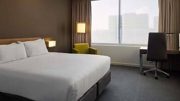 Een hotelkamer met een groot bed, een bureau, een stoel, een lamp en een raam met uitzicht op gebouwen.
