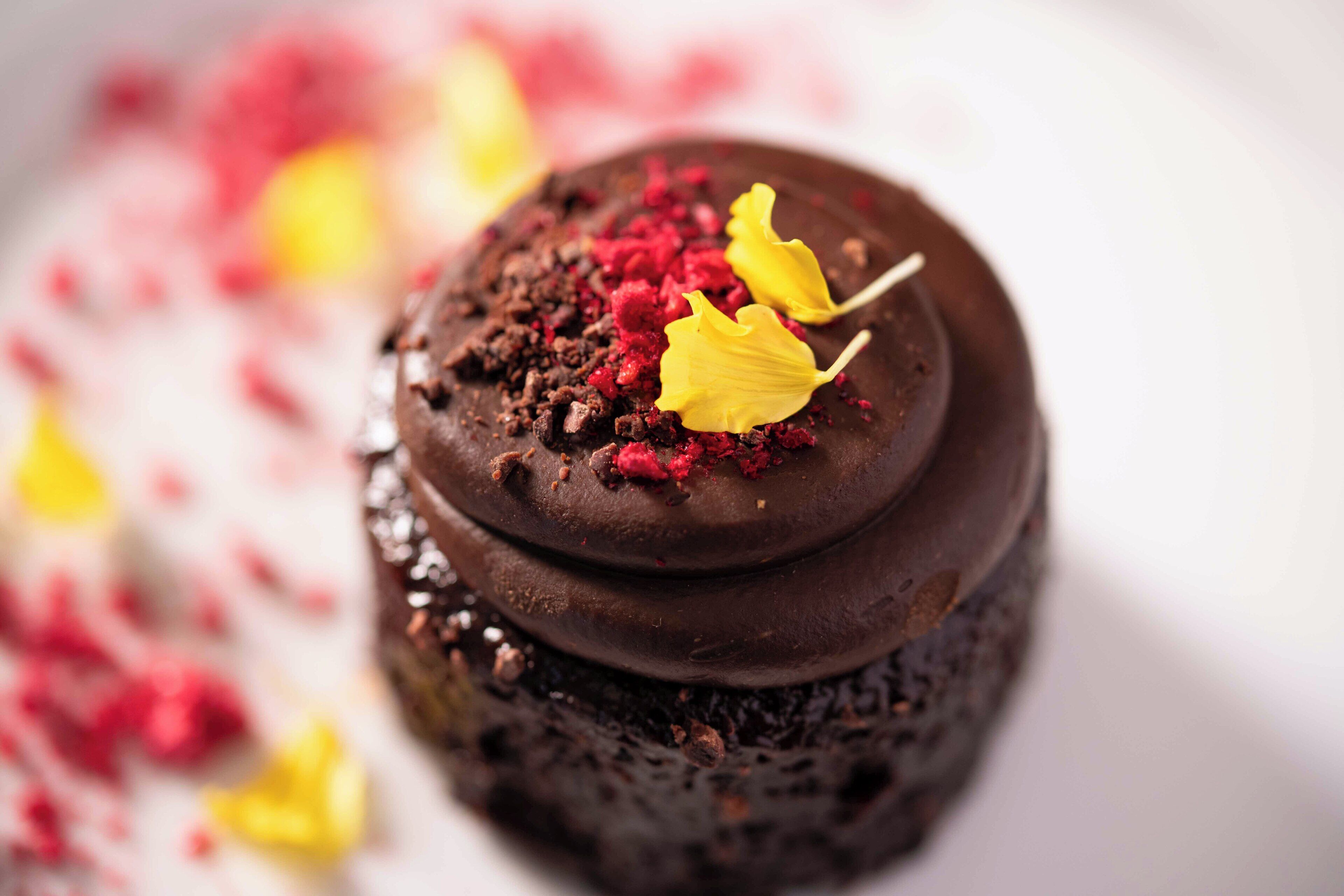 Postre de xocolata amb acabat brillant, coronat amb molles vermelles i flors comestibles grogues.