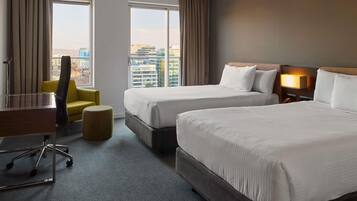 A hotel room with two beds, a desk, a chair, and a view of the cityscape.