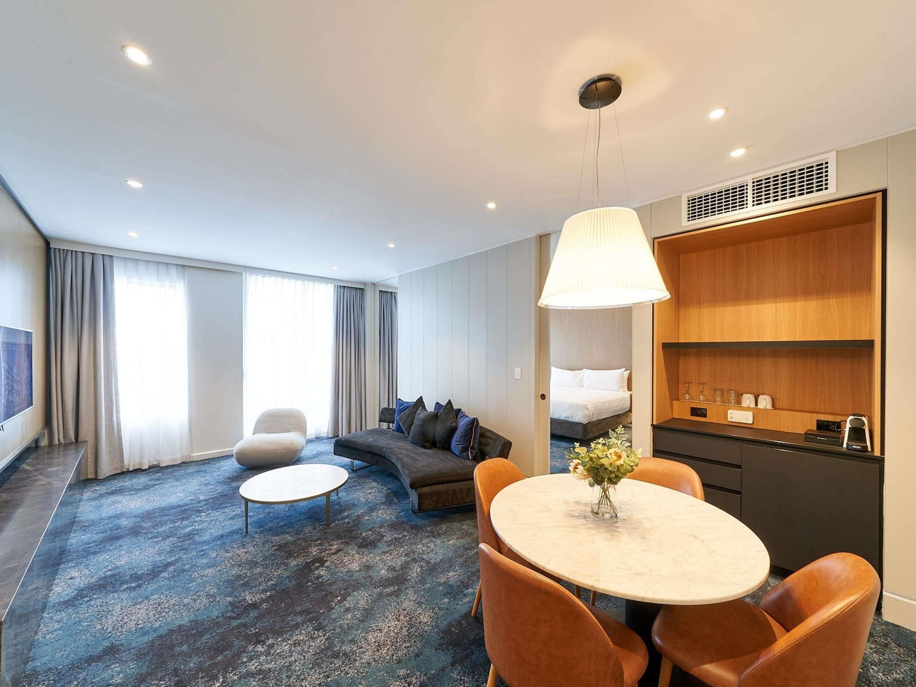 A modern hotel room with a sofa, a dining table with chairs, a kitchen area, and a large window with curtains.