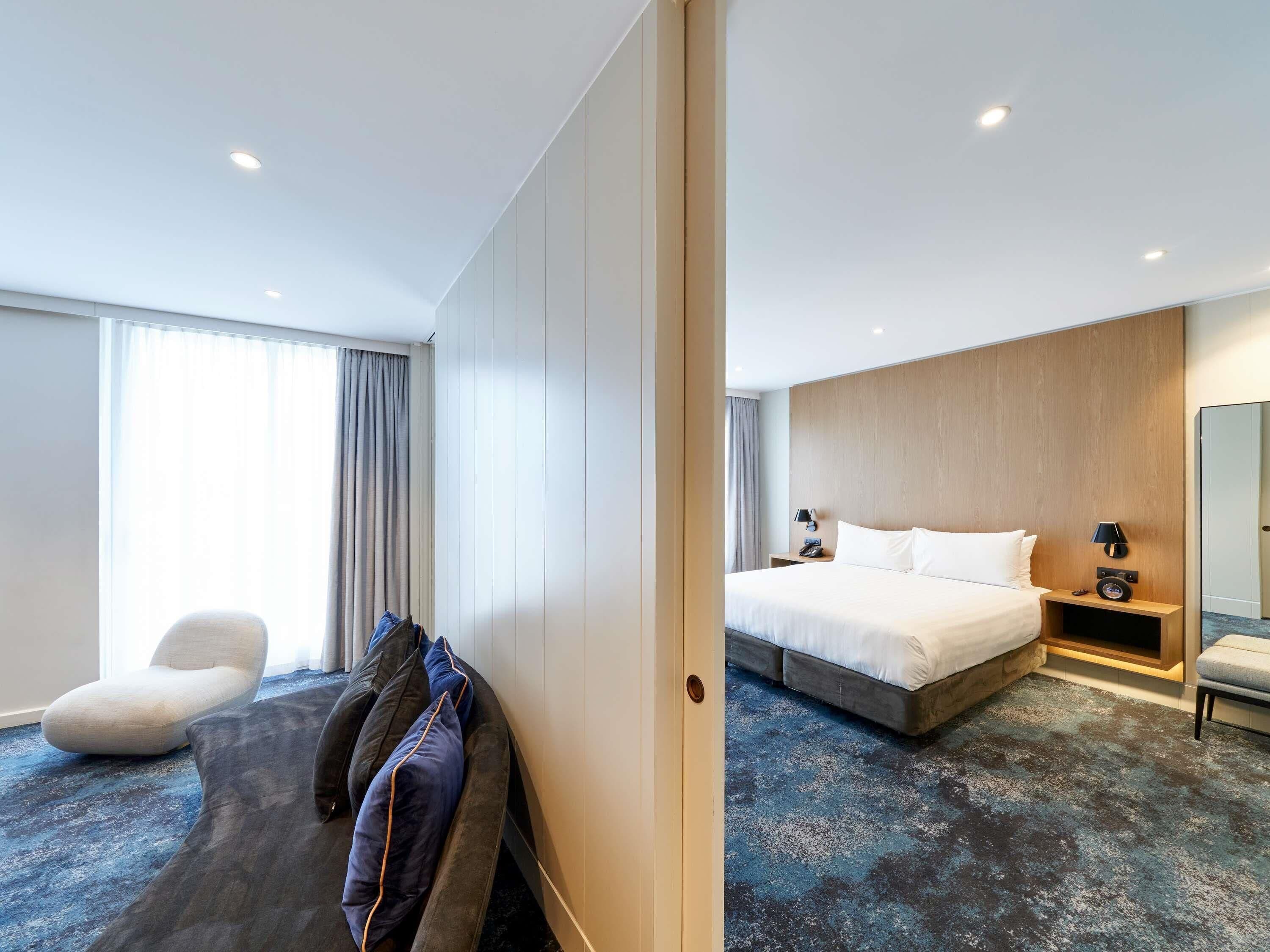 A hotel room with a bed, bedside tables, a chair, a sofa, and a large window with curtains.