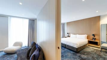 Habitación de hotel con cama, mesitas de noche, una silla, sofá y un amplio ventanal con cortinas.