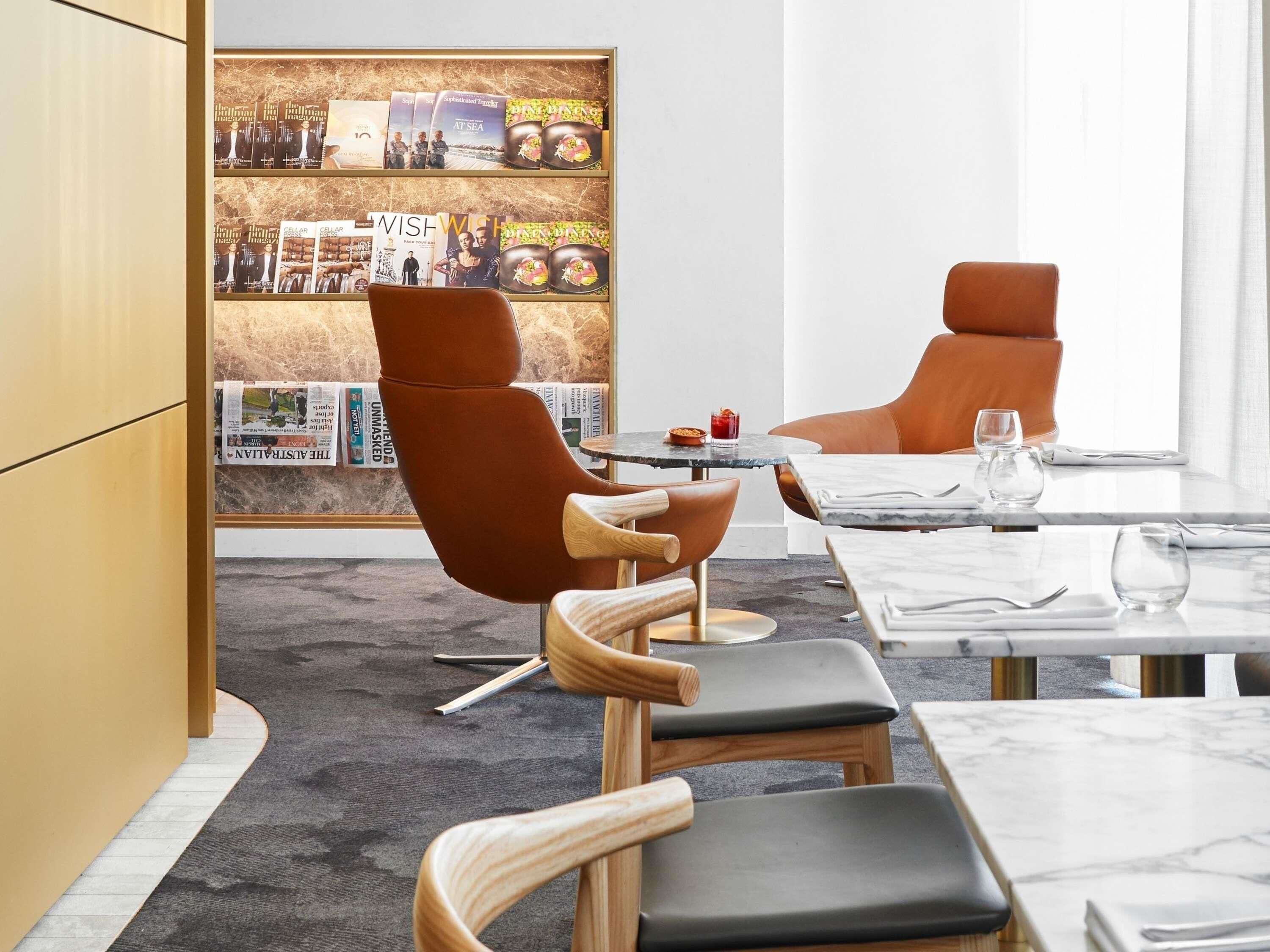 A modern hotel room with a marble table, leather chairs, a bookshelf with magazines, and a large window.