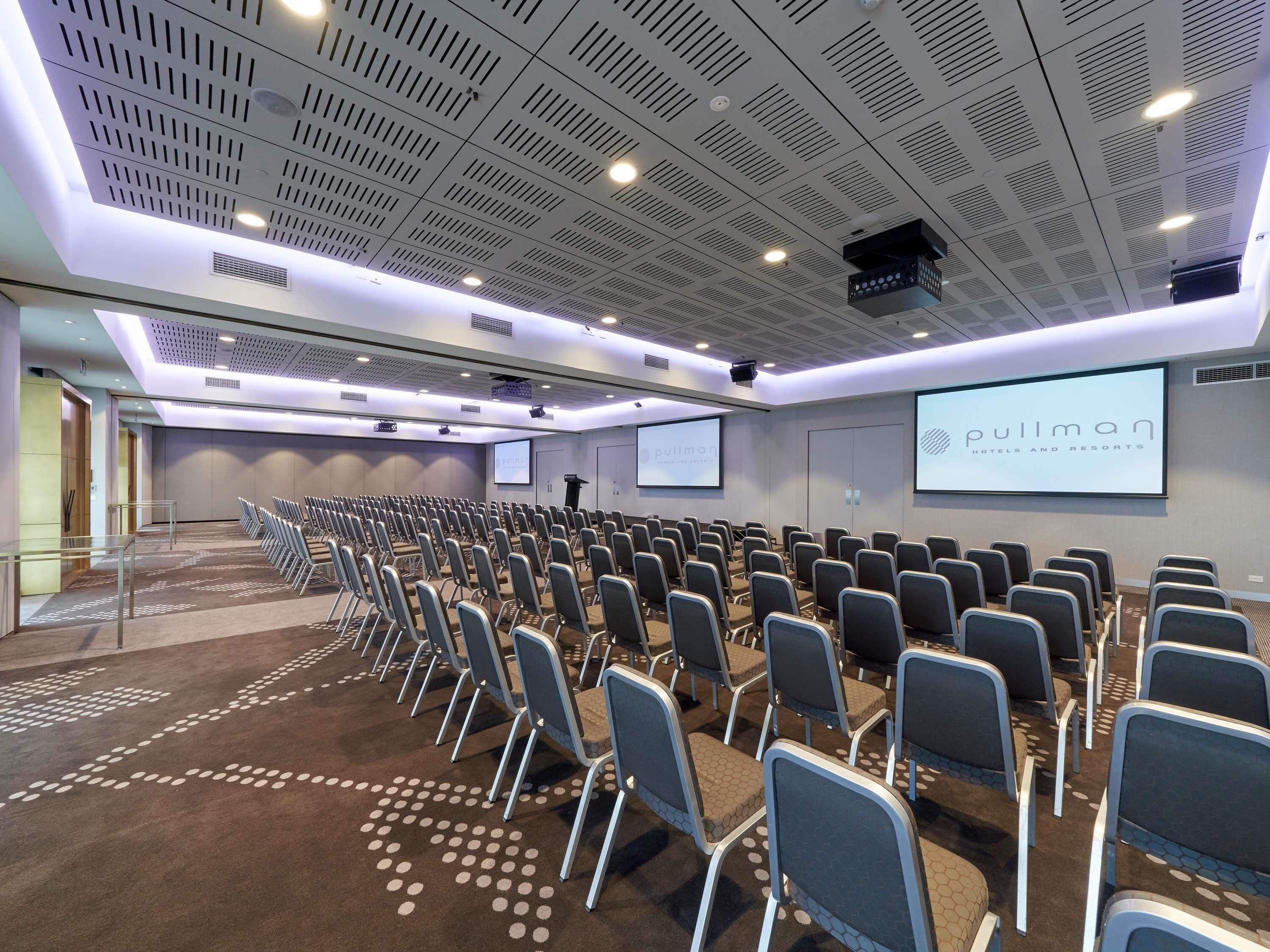 A conference room with rows of chairs, a projector screen, and a 'pullman' branding on the wall.