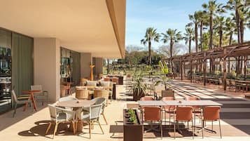 Un patio al aire libre con diferentes disposiciones de asientos, incluyendo mesas y sillas, rodeado de macetas con plantas y palmeras.