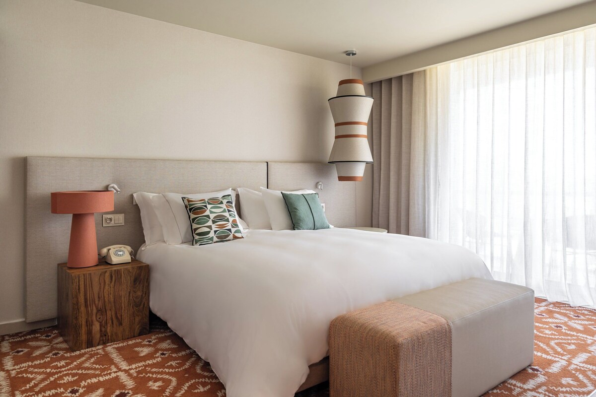 A hotel room with a large bed, a bedside lamp, a telephone, and a window with curtains.