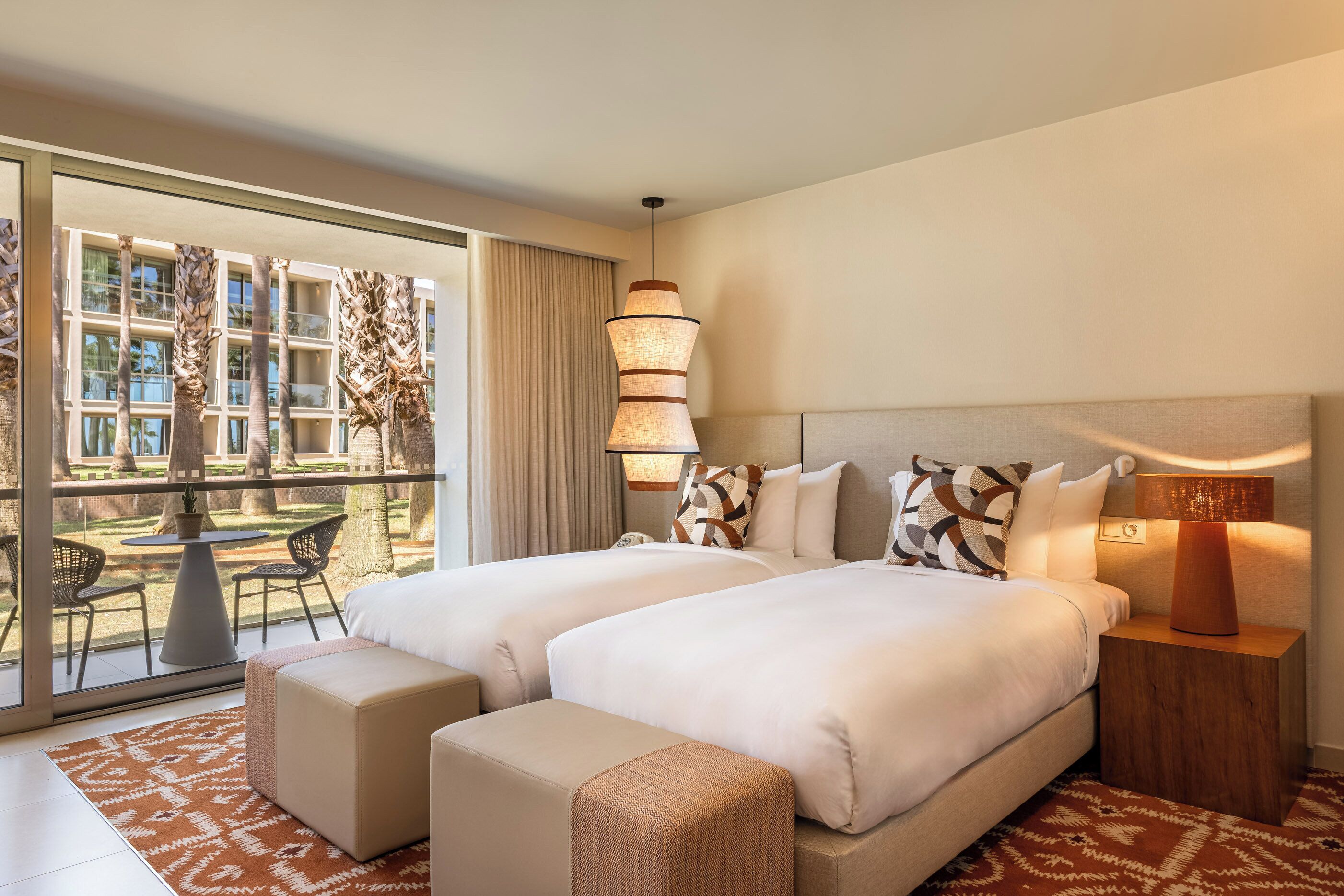 A hotel room with two beds, a balcony with a table and chairs, and a view of palm trees.