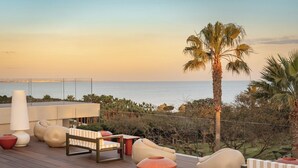 A deck with modern seating, a large white lamp, and a view of the ocean at sunset.