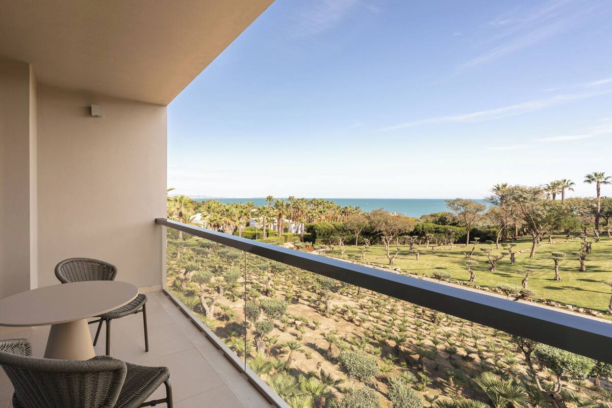 A balcony with wicker chairs and a table, overlooking a landscaped garden and a distant view of the sea.
