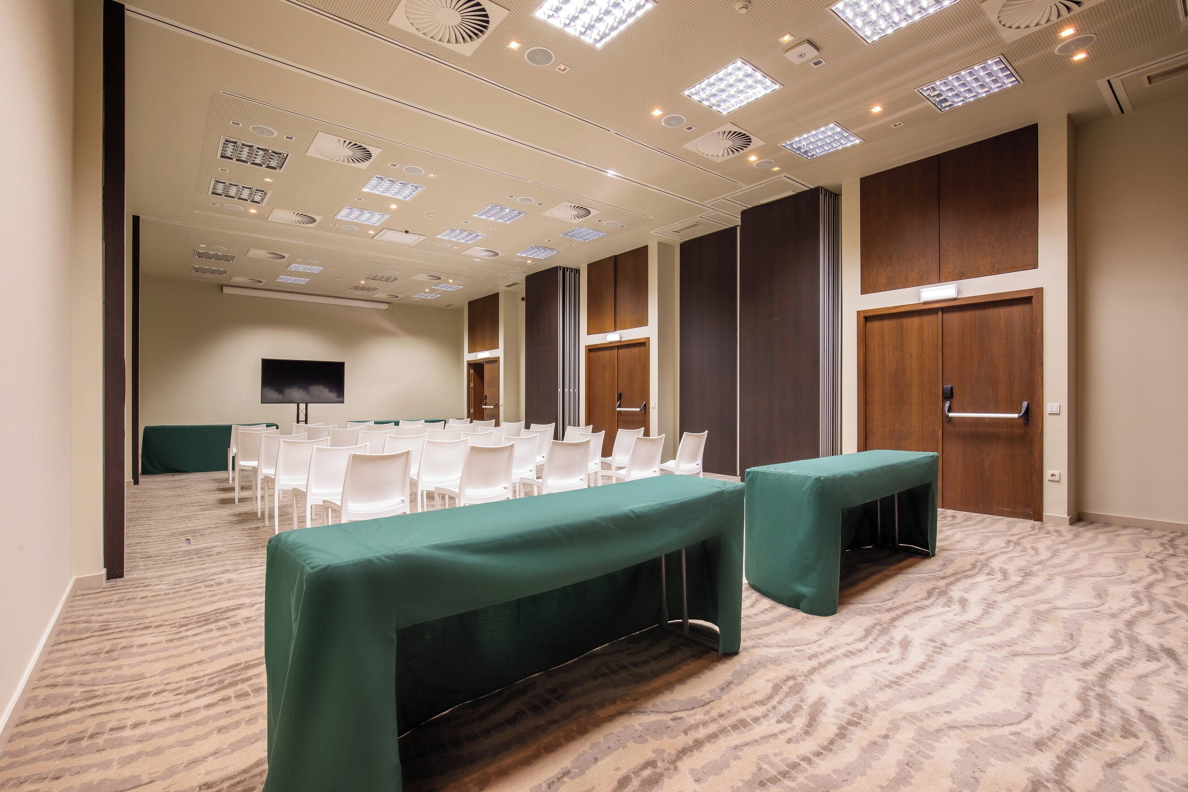 A conference room with a long table, white chairs, and a green tablecloth. There are wooden doors and a television on the wall.