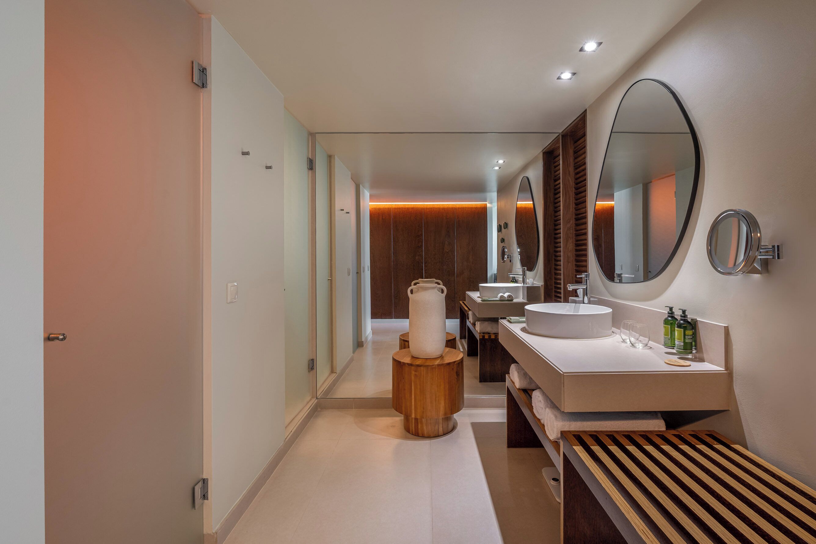 A modern bathroom with a large mirror, two sinks, and a wooden bench.