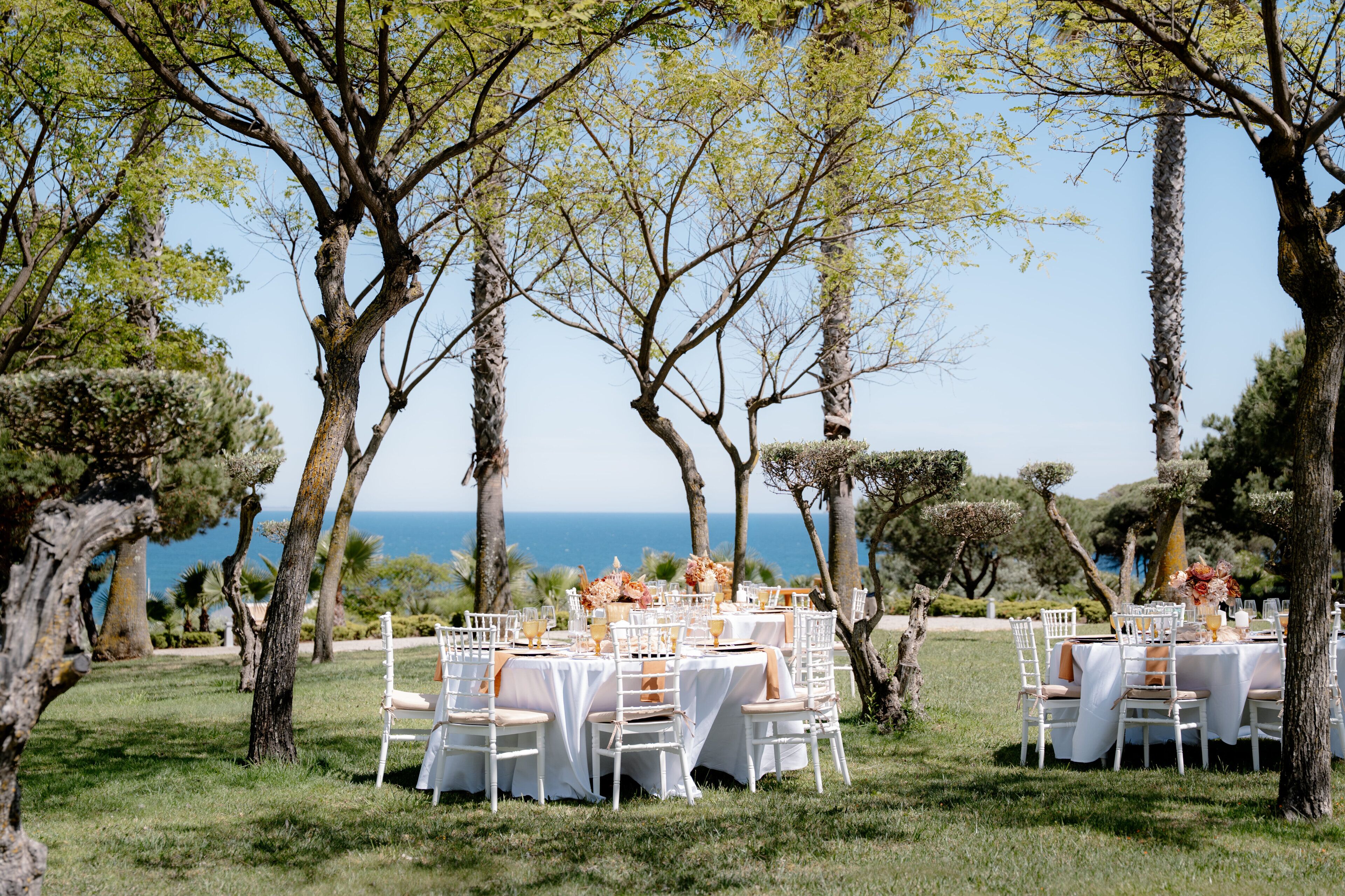 An outdoor dining area with tables set for guests, surrounded by trees and overlooking a body of water.