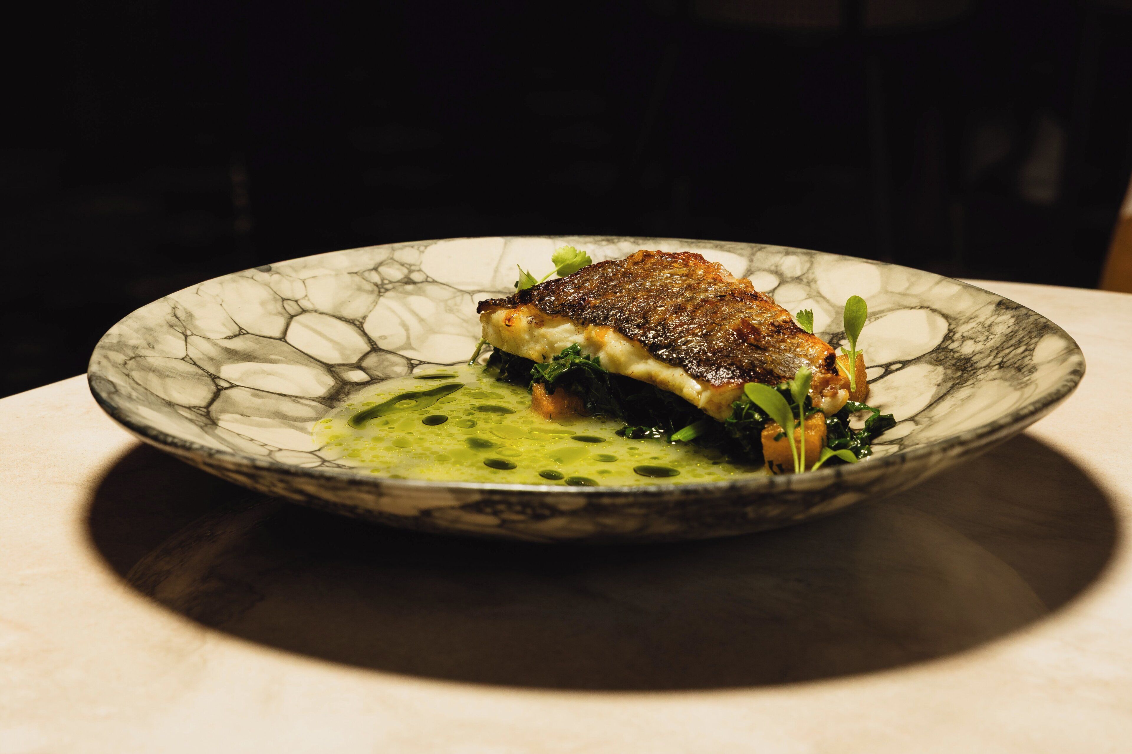 A plate with a piece of food, possibly a steak, on a textured plate with a green sauce and greens.