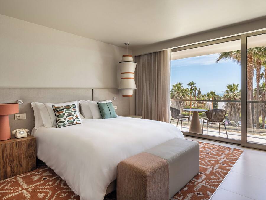 Une chambre d’hôtel moderne dotée d’un grand lit, d’une table de chevet avec une lampe, d’un téléphone et offrant une vue sur les palmiers et l’océan grâce à de grandes fenêtres.
