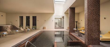 A modern indoor swimming pool with lounge chairs and a skylight.