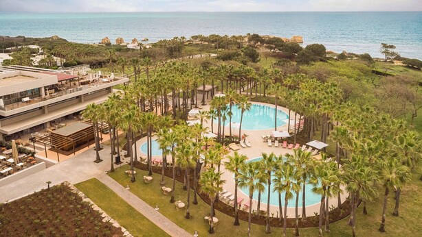 A resort with a large circular pool surrounded by palm trees, overlooking the sea.