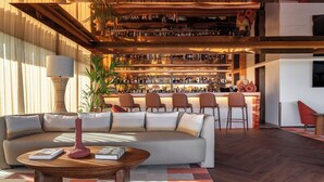 A modern bar area with a sofa and chairs in the foreground, a bar counter with numerous bottles, and a large plant.
