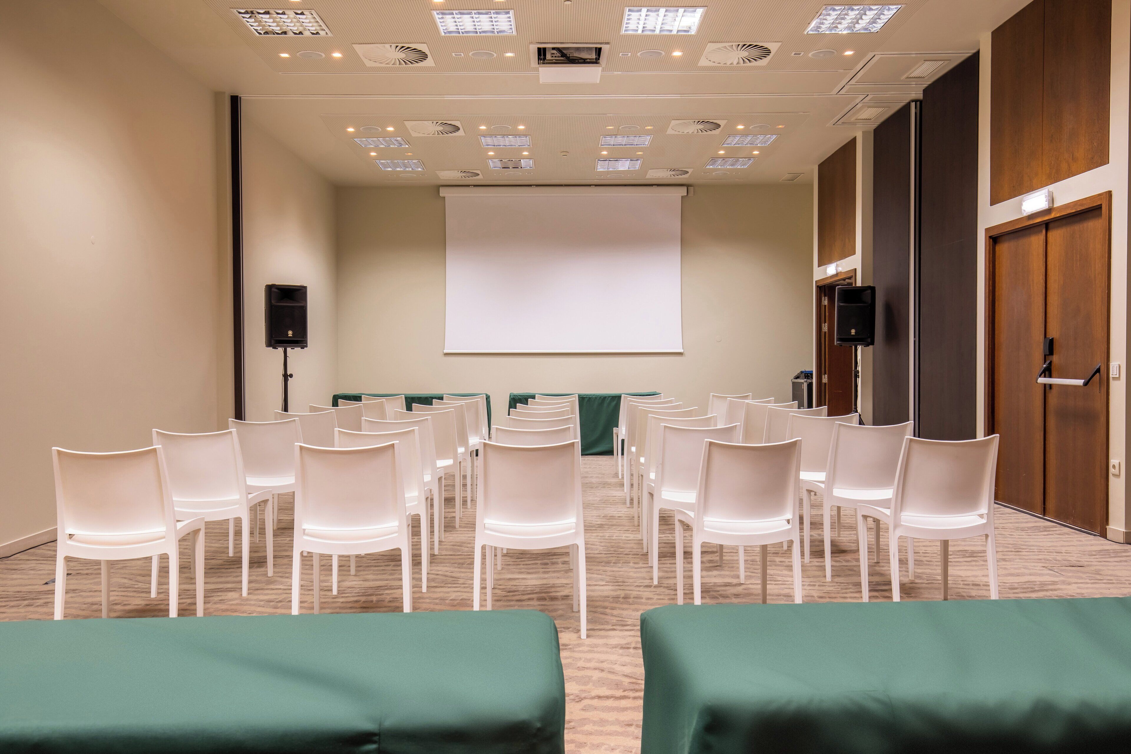 Uma sala de conferências com fileiras de cadeiras brancas, uma tela de projeção grande e piso carpeteado.