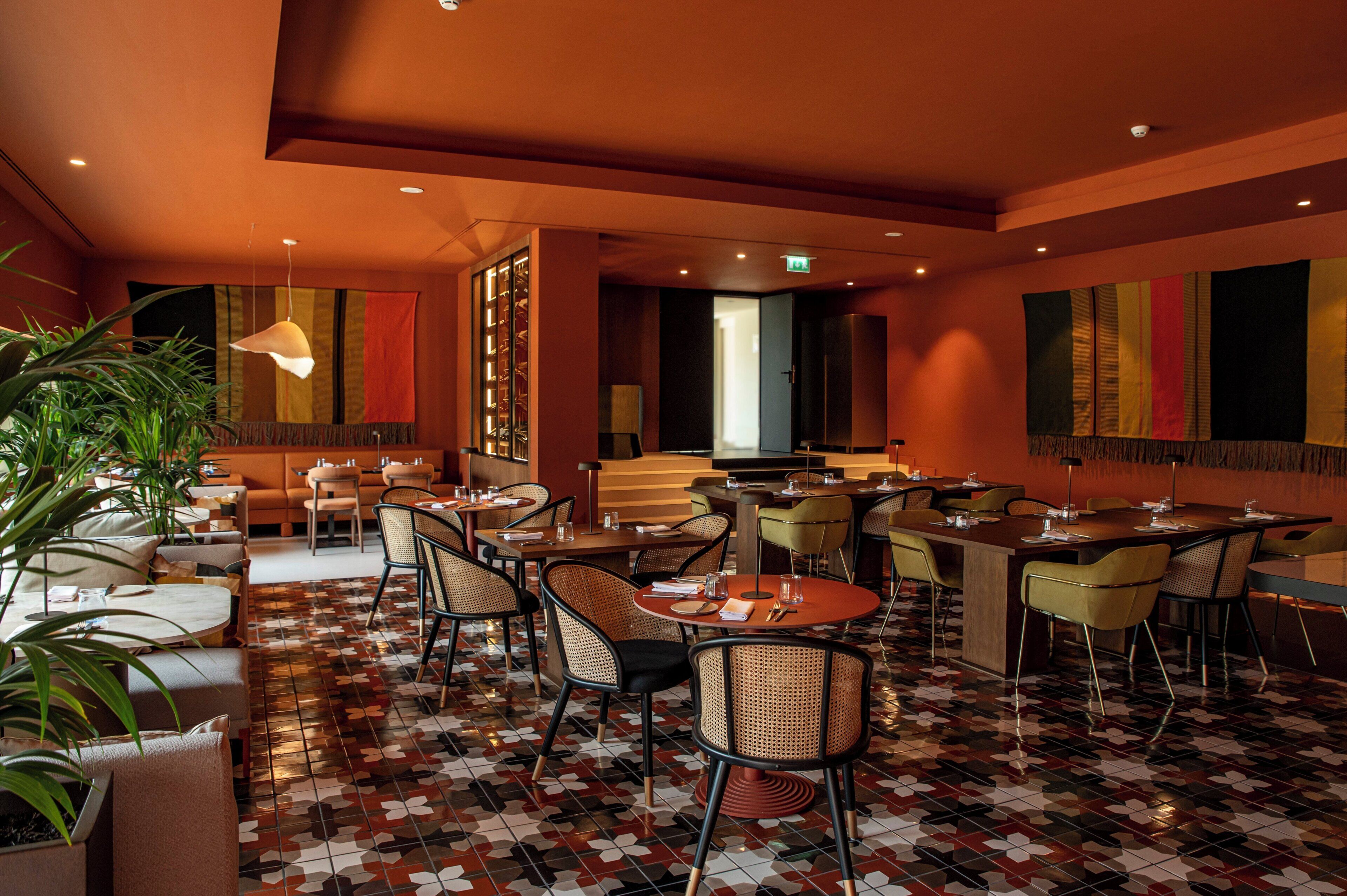 A restaurant with a modern interior, featuring a mix of round and rectangular tables, wicker chairs, and a patterned floor.