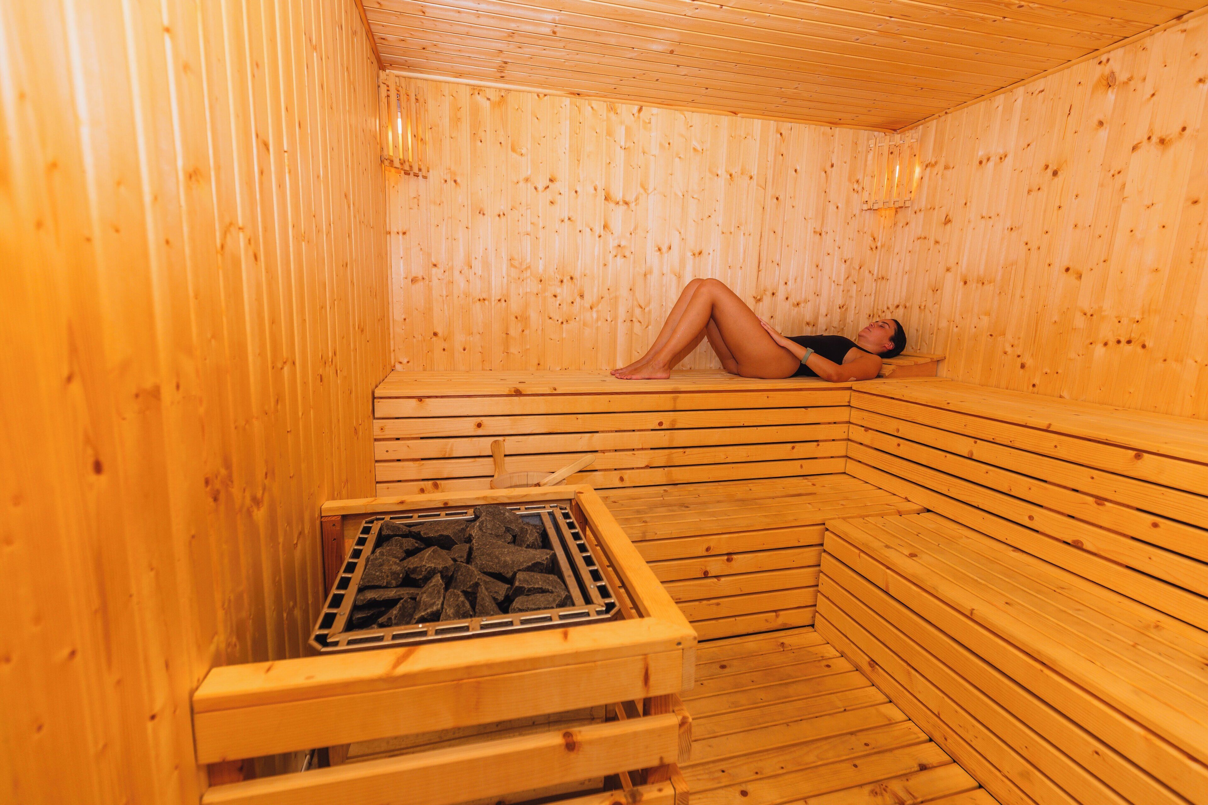 A sauna with wooden walls and benches, a person lying on a bench, and a grate with stones.