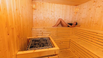 A sauna with wooden walls and benches, a person lying on a bench, and a grate with stones.
