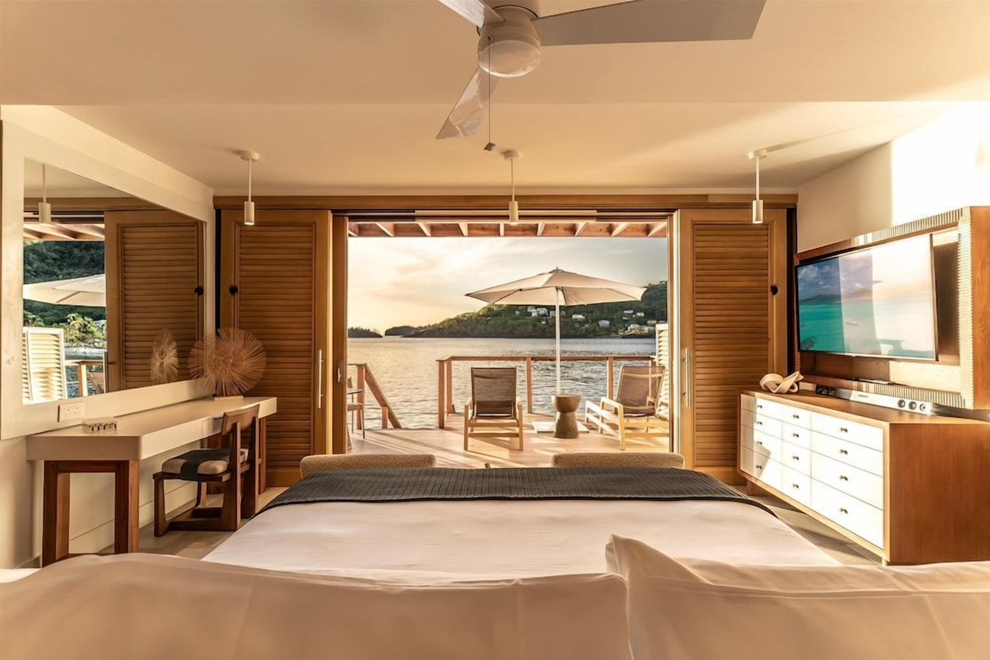 A bedroom with a large bed, a TV, a desk, and a view of a body of water.