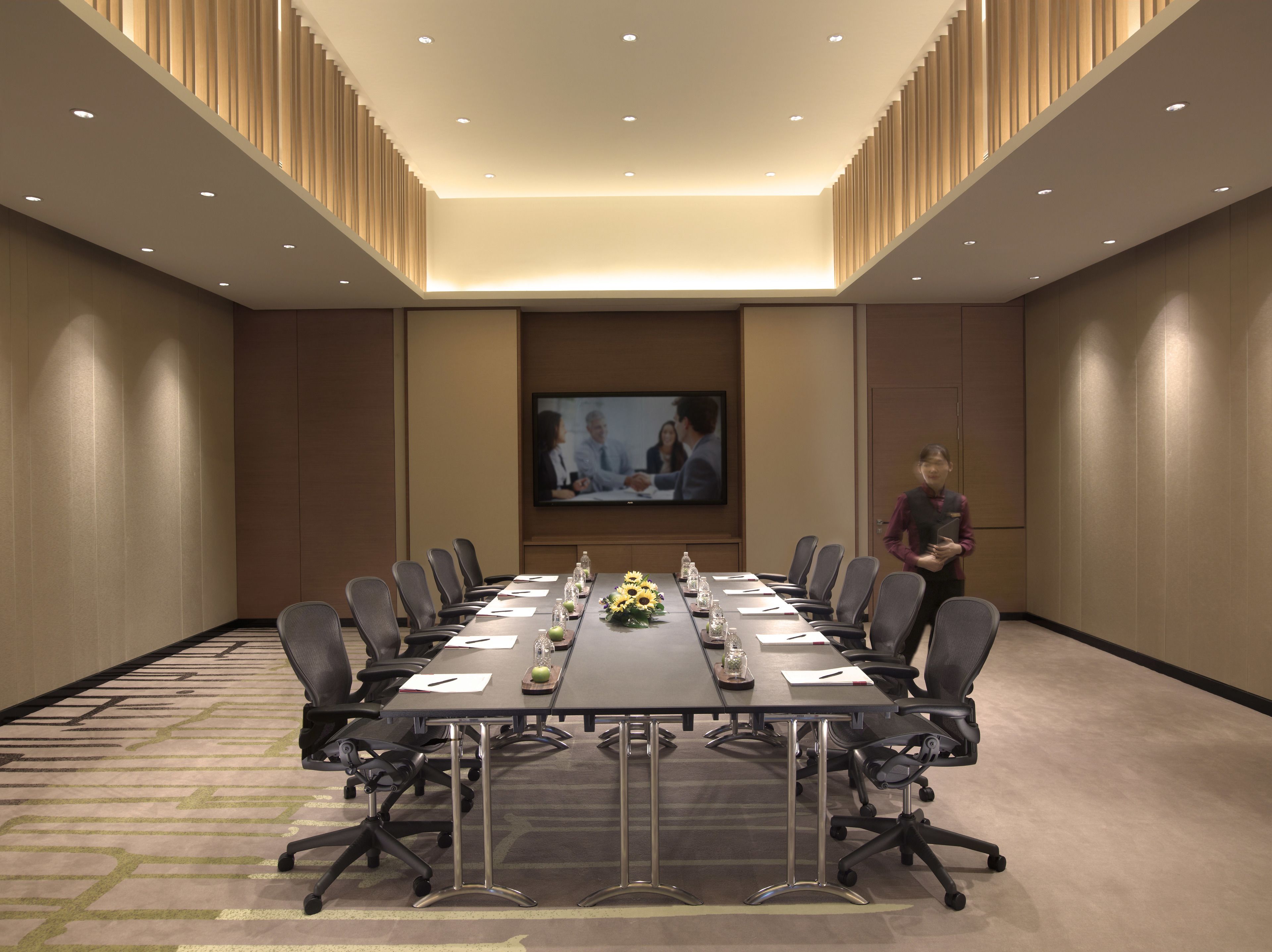 A conference room with a long table, chairs, a TV screen, and a person standing by the table.