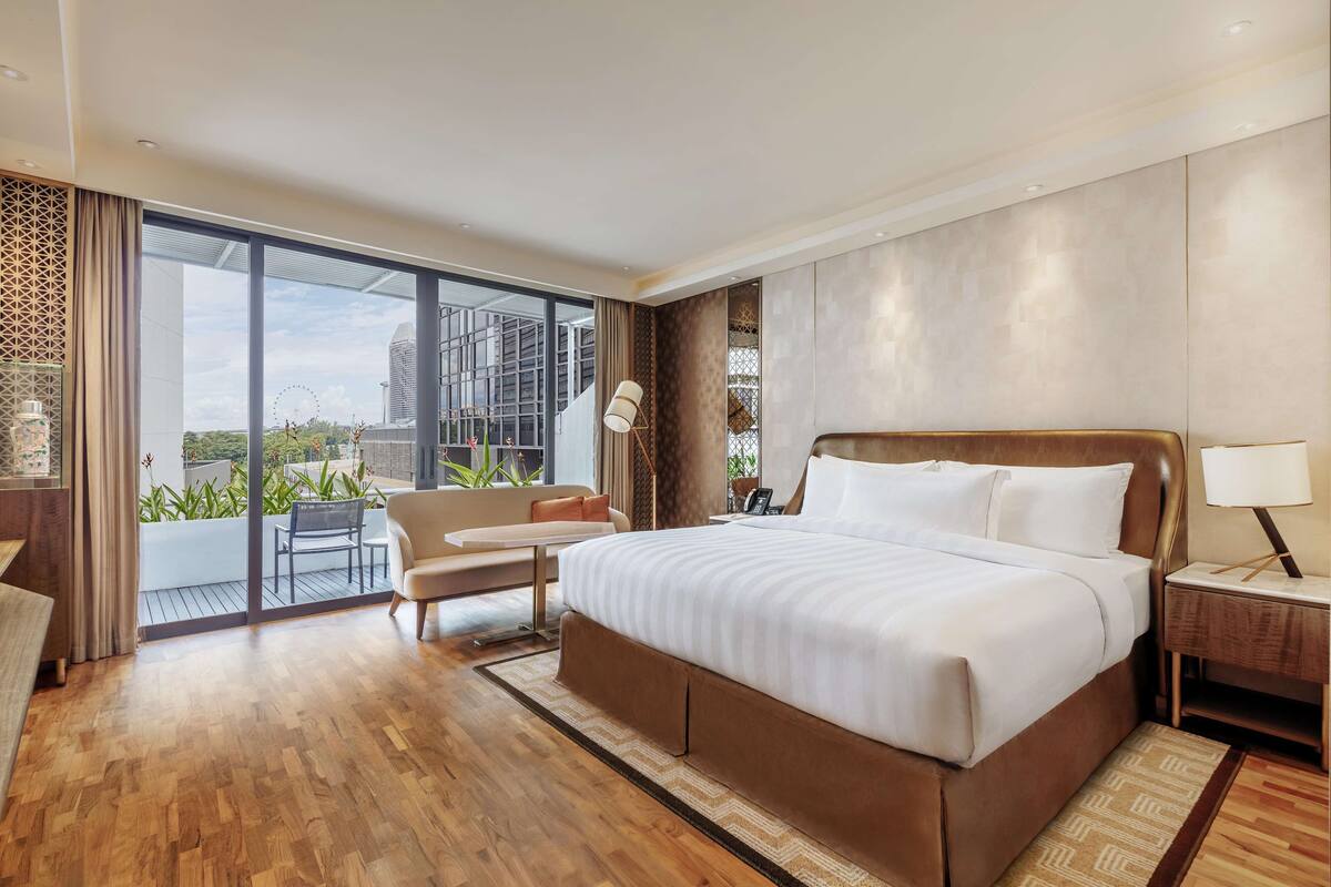Quarto de hotel moderno com uma cama grande, área de estar com sofá e cadeiras, varanda com plantas e vista para a cidade.