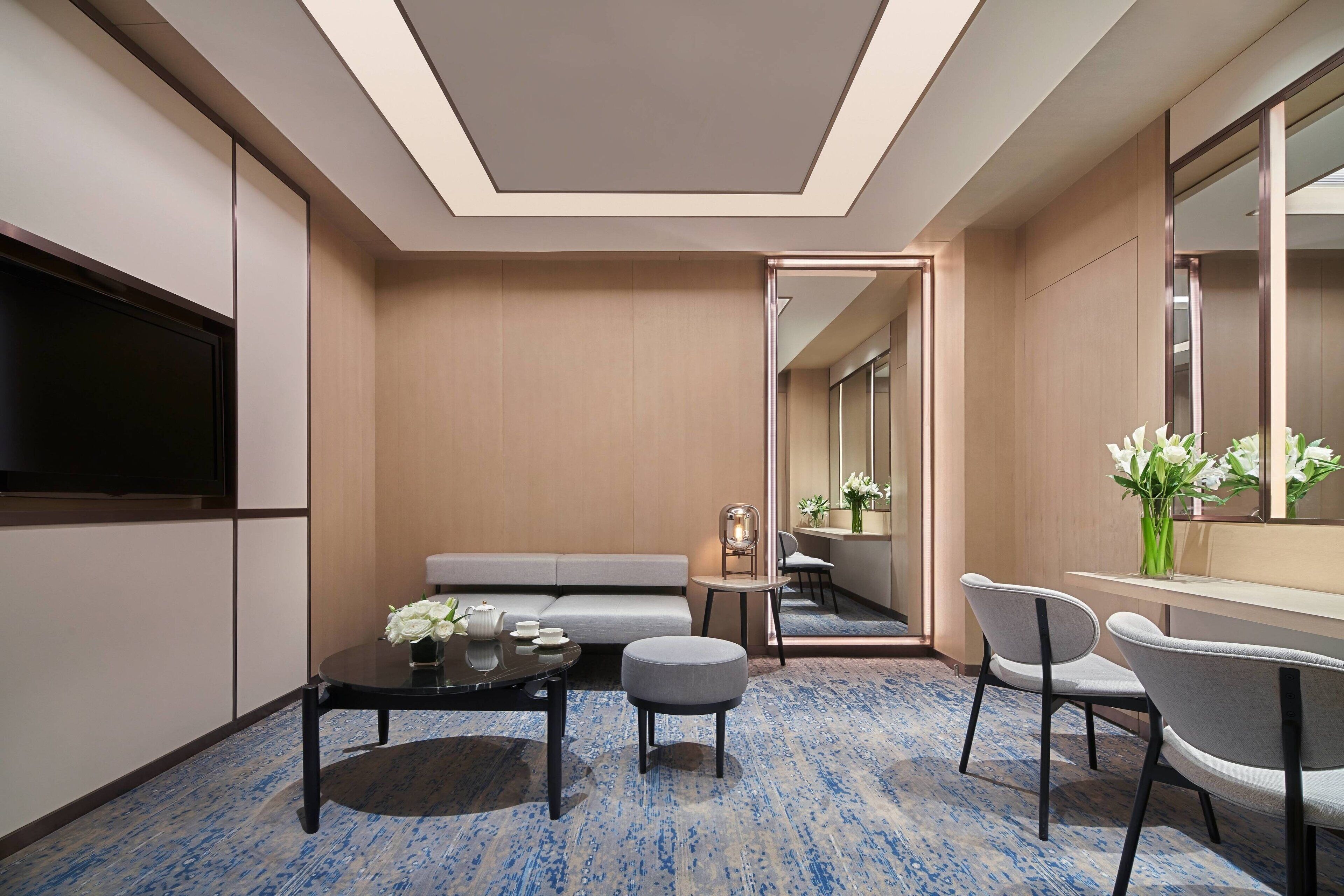 A modern hotel room with a sofa, a small round table with a vase of flowers, a chair, and a flat-screen TV mounted on the wall.