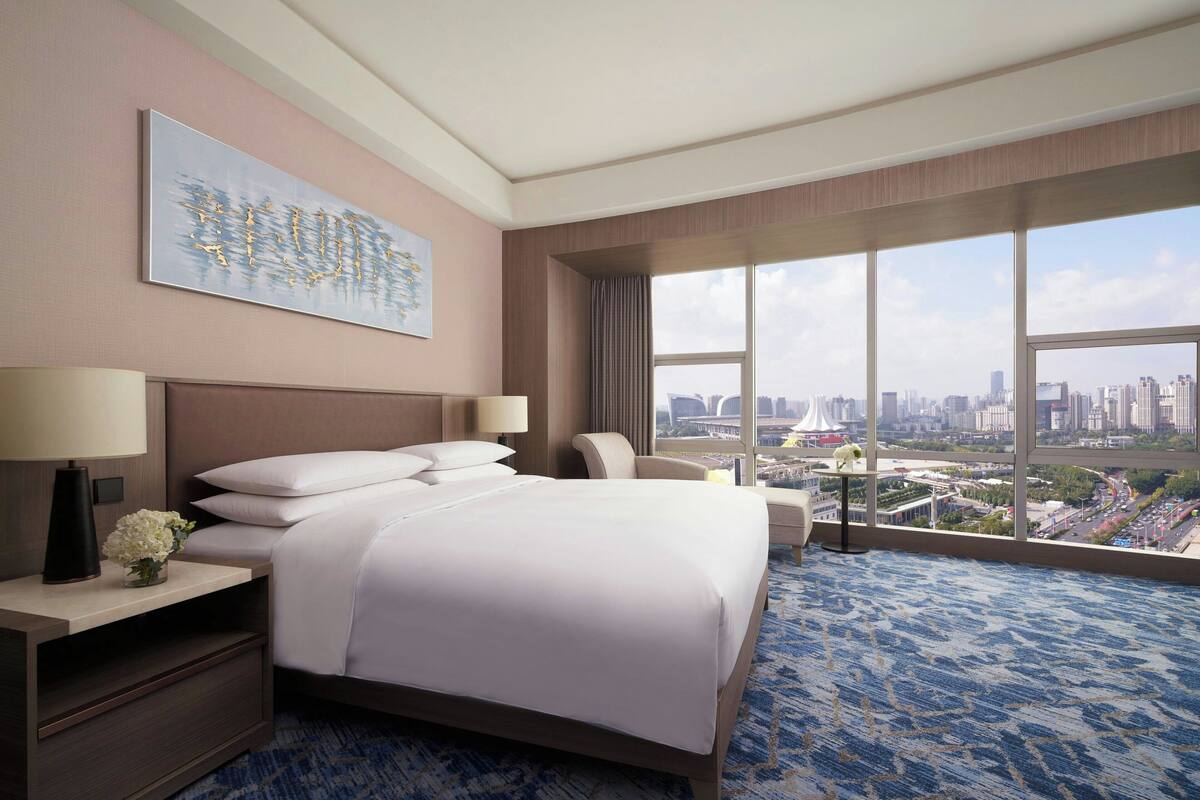 Habitación de hotel con una cama grande, un escritorio con lámpara, una silla y un ventanal con vistas a la ciudad.