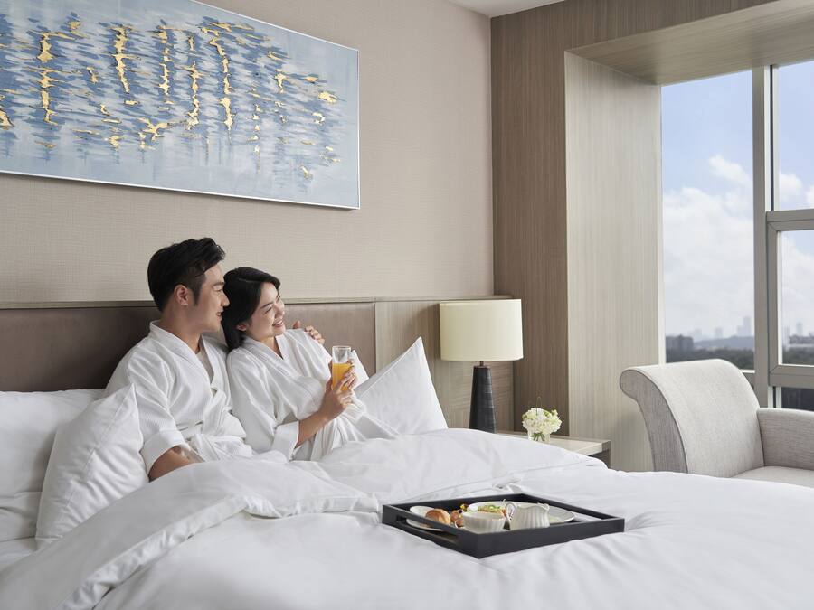 A hotel room with a large window, a bed with white linens, a tray with breakfast items, and a painting on the wall.