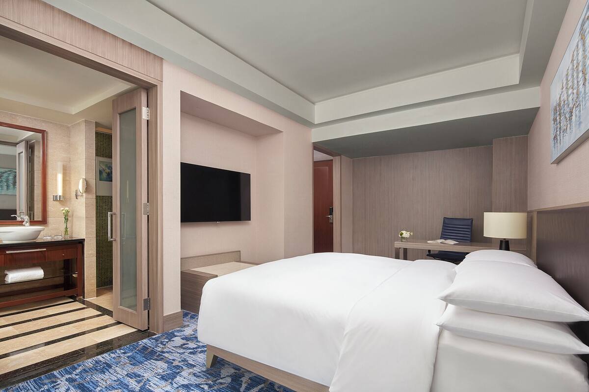 Una habitación de hotel con una cama grande, un escritorio con una silla, un televisor montado en la pared y un baño con lavamanos y espejo.