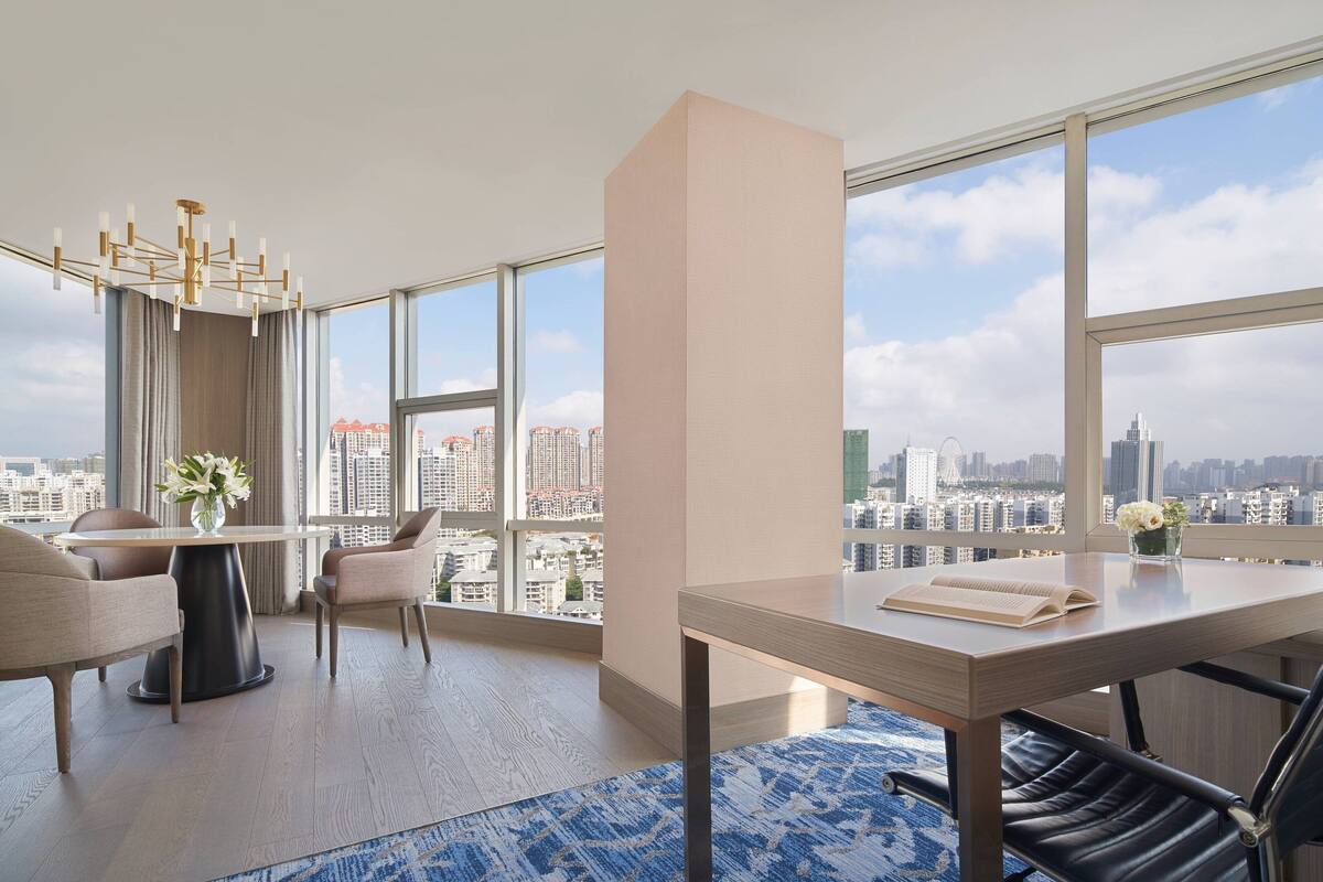 Una habitación de hotel moderna con un amplio ventanal que ofrece vistas a la ciudad, una mesa redonda, dos sillas, un florero con flores y un accesorio de iluminación decorativo.