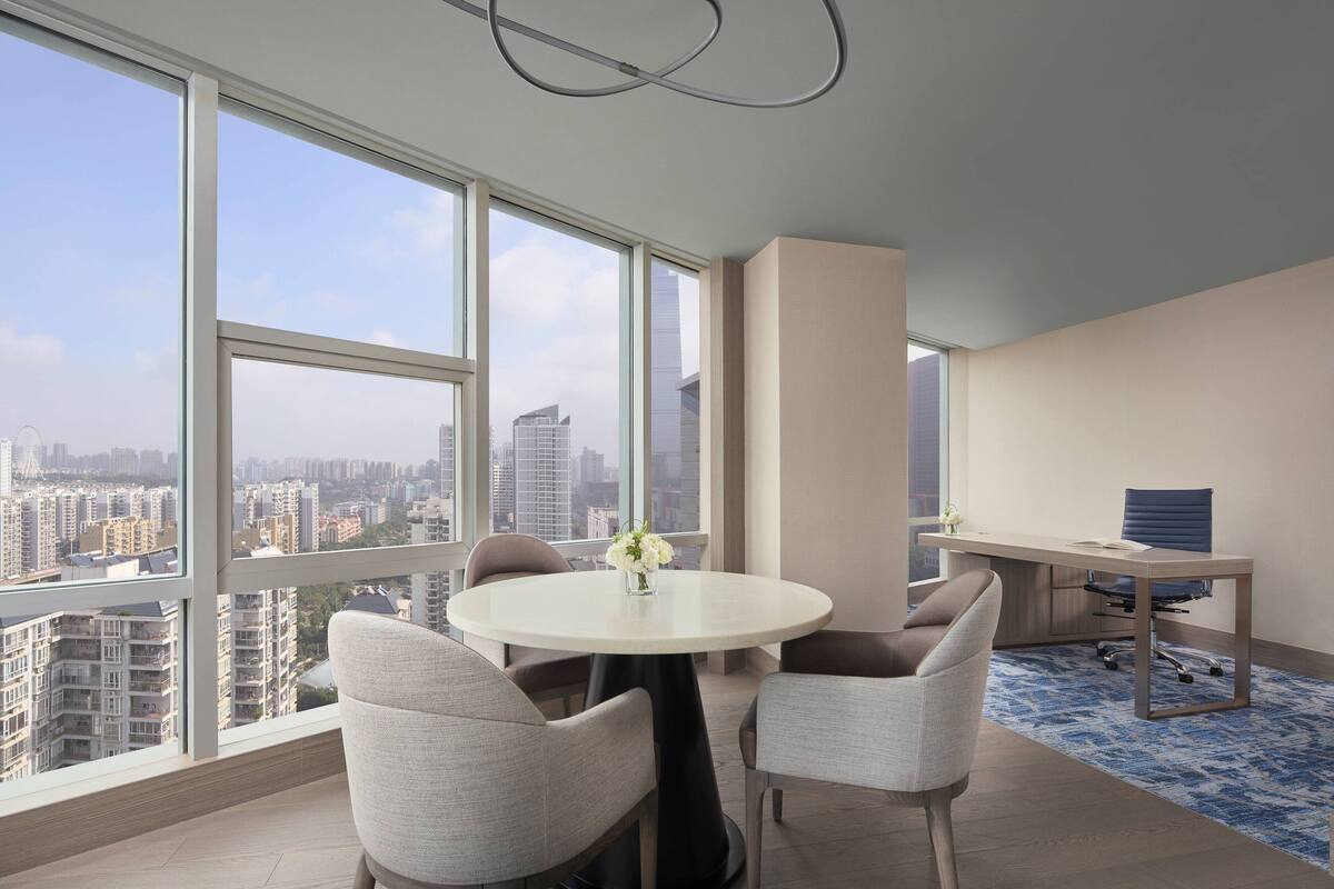 Un espacio de oficina moderno con una mesa redonda, dos sillas, un escritorio con una silla y ventanas amplias que ofrecen vistas a la ciudad.