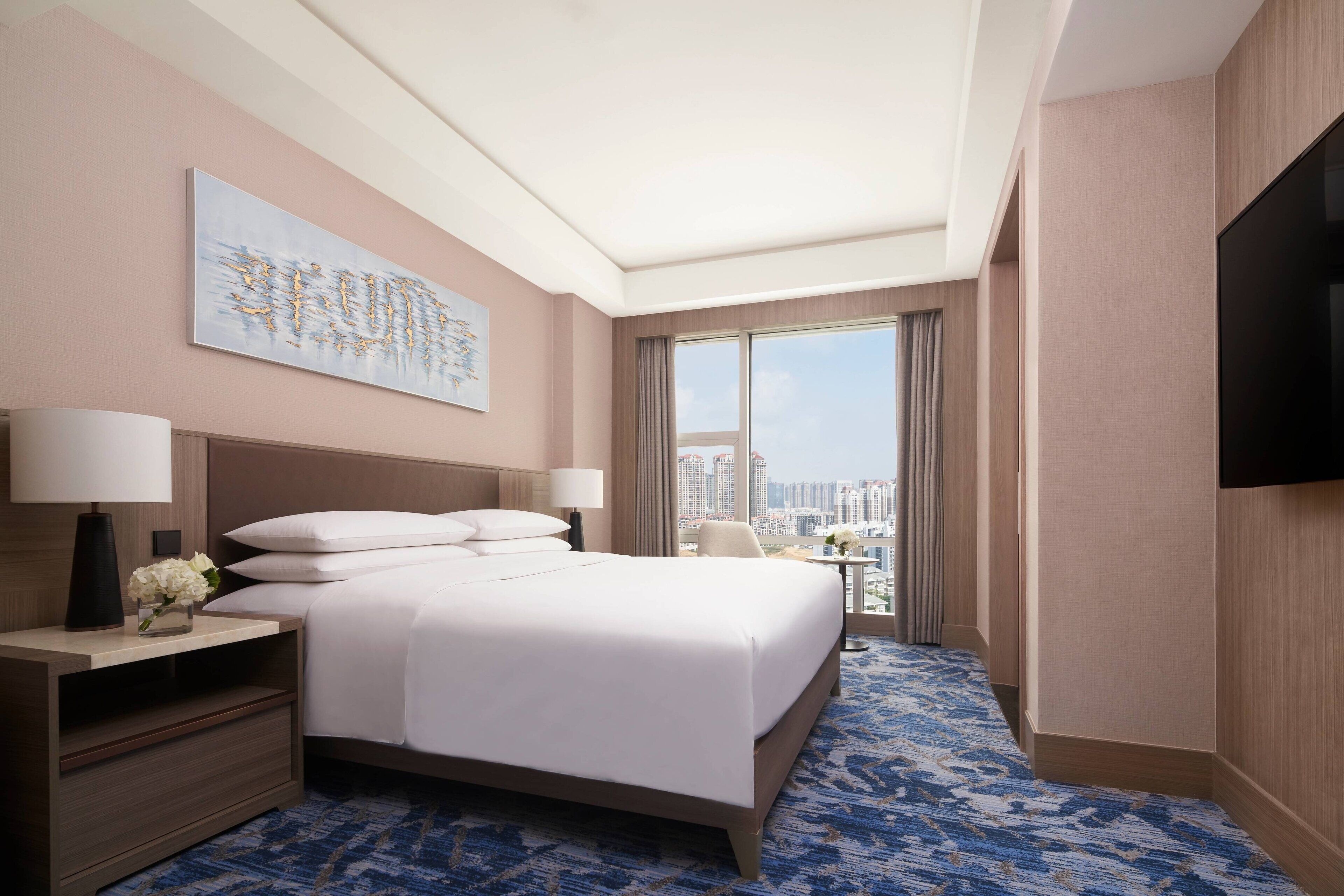 A hotel room with a large bed, a flat-screen TV, a nightstand with a lamp, and a view of the city through a large window.