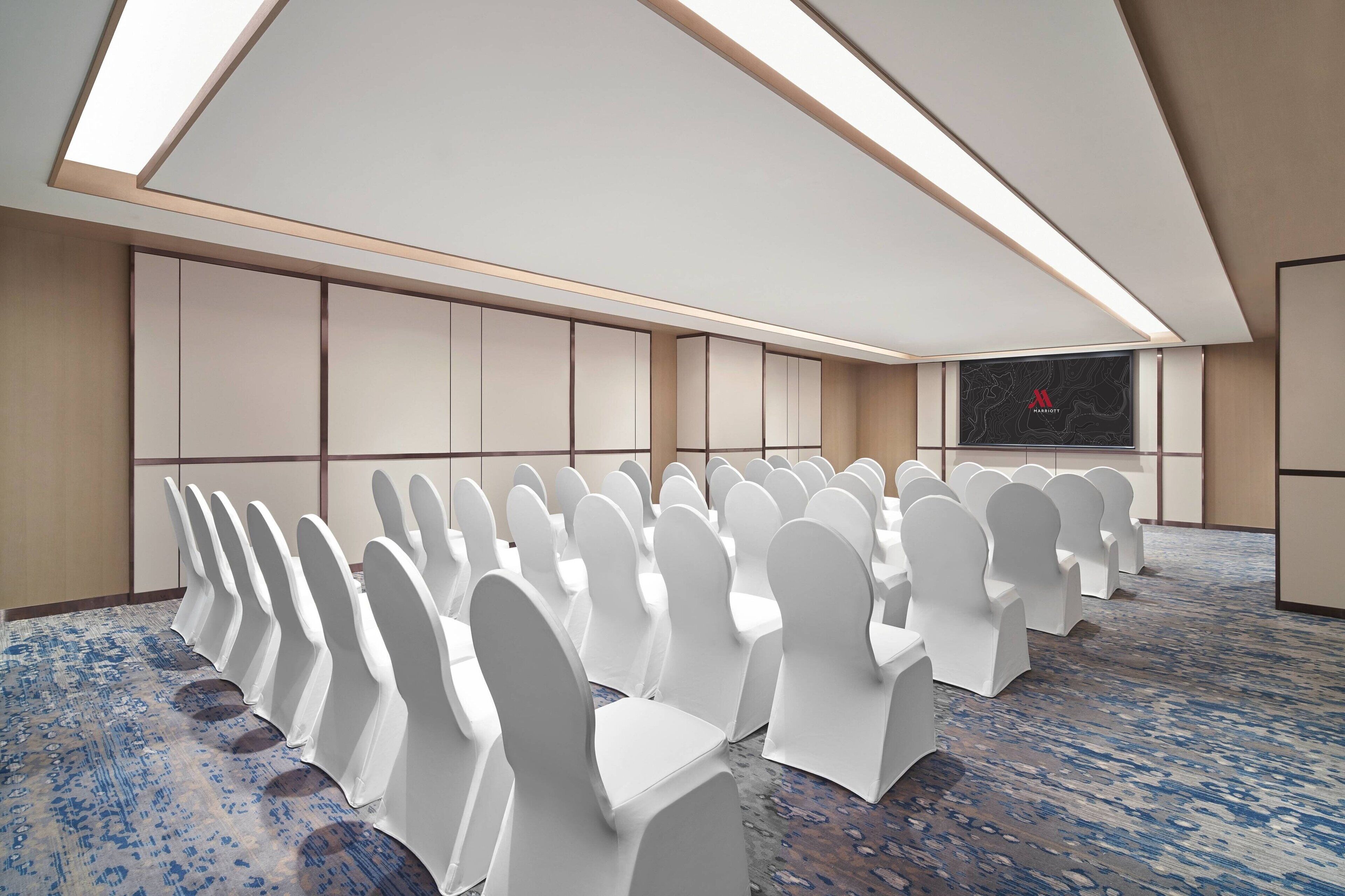 Un salón de conferencias con hileras de sillas blancas, suelo alfombrado y una pantalla grande en la pared.