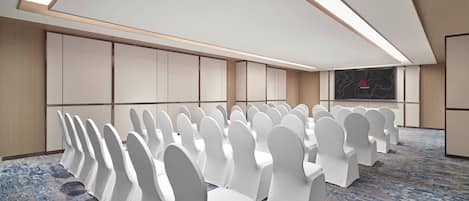 Un salón de conferencias con hileras de sillas blancas, suelo alfombrado y una pantalla grande en la pared.