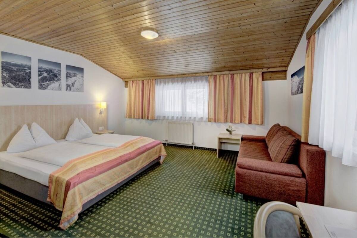 Une chambre d’hôtel avec un plafond en bois, un lit recouvert d’une courtepointe à rayures, un canapé rouge, une petite table et une fenêtre avec des rideaux.