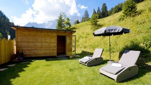 A wooden cabin with an open door, two lounge chairs, and an umbrella on a grassy lawn with a mountainous backdrop. - Hotel-Restaurant Bike&Snow Lederer (Muehlbach Am Hochkoenig)