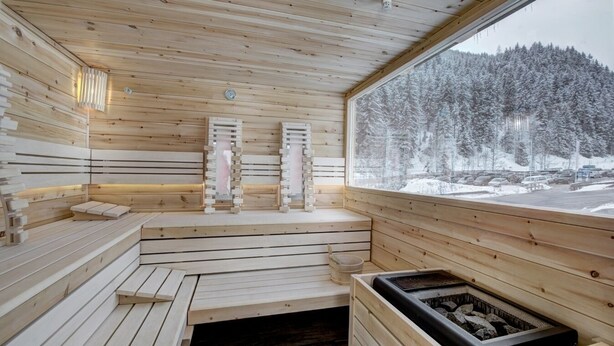 A wooden sauna with benches and a sauna heater, a window with a snowy forest view, and a wall-mounted heater. - Hotel-Restaurant Bike&Snow Lederer (Muehlbach Am Hochkoenig)