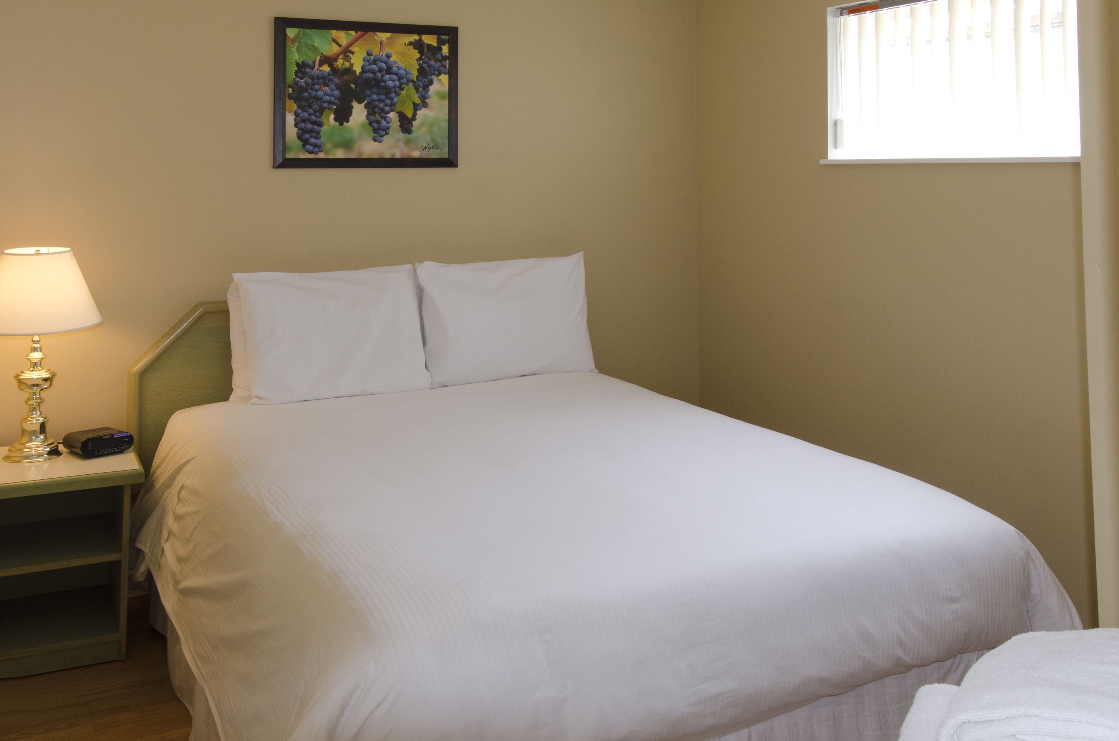 A neatly made bed with white linens, a bedside lamp, and a wall-mounted painting of grapes.