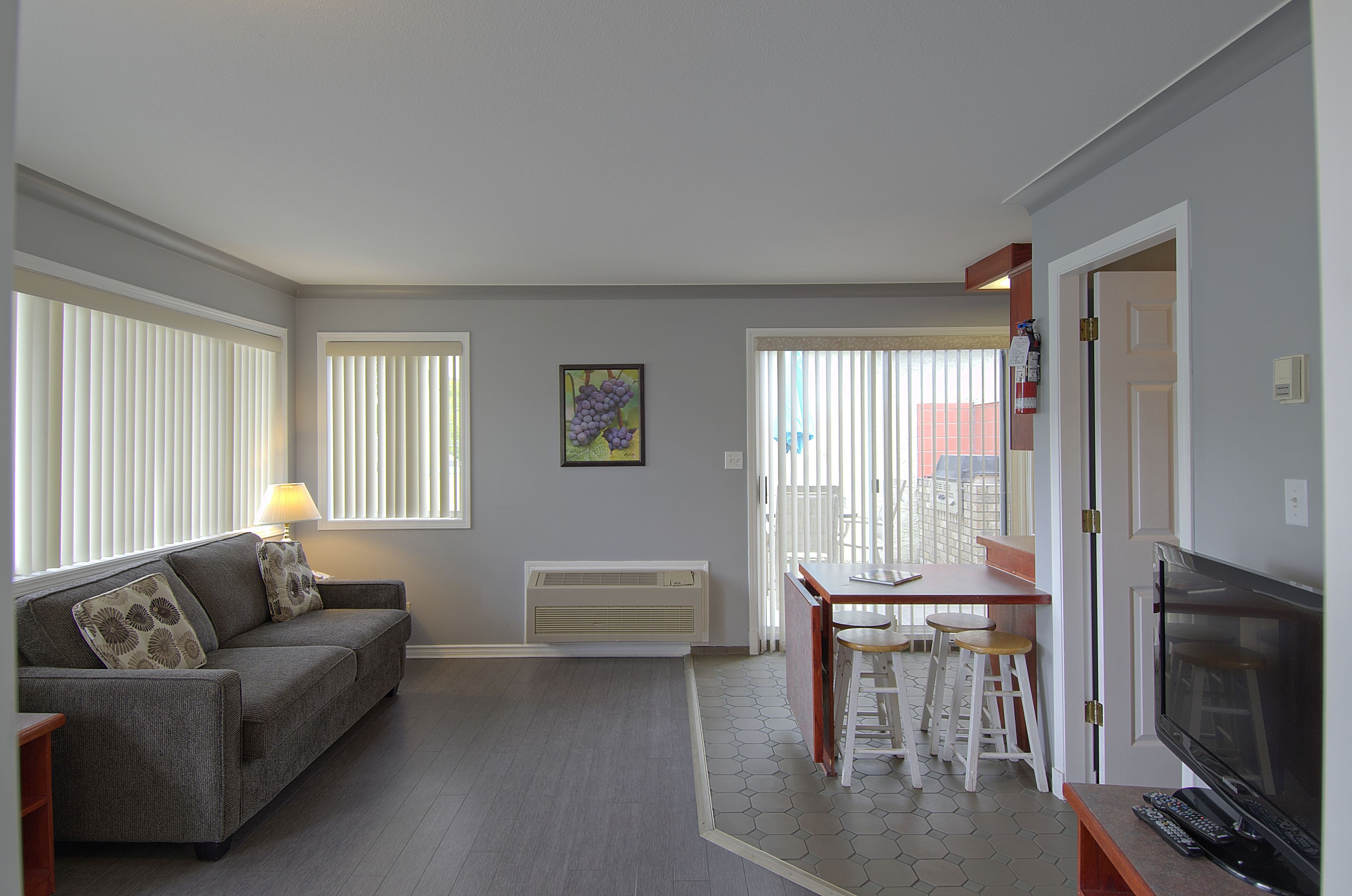 A living room with a sofa, a table, and a TV. There is a window with blinds, a picture on the wall, and a radiator.