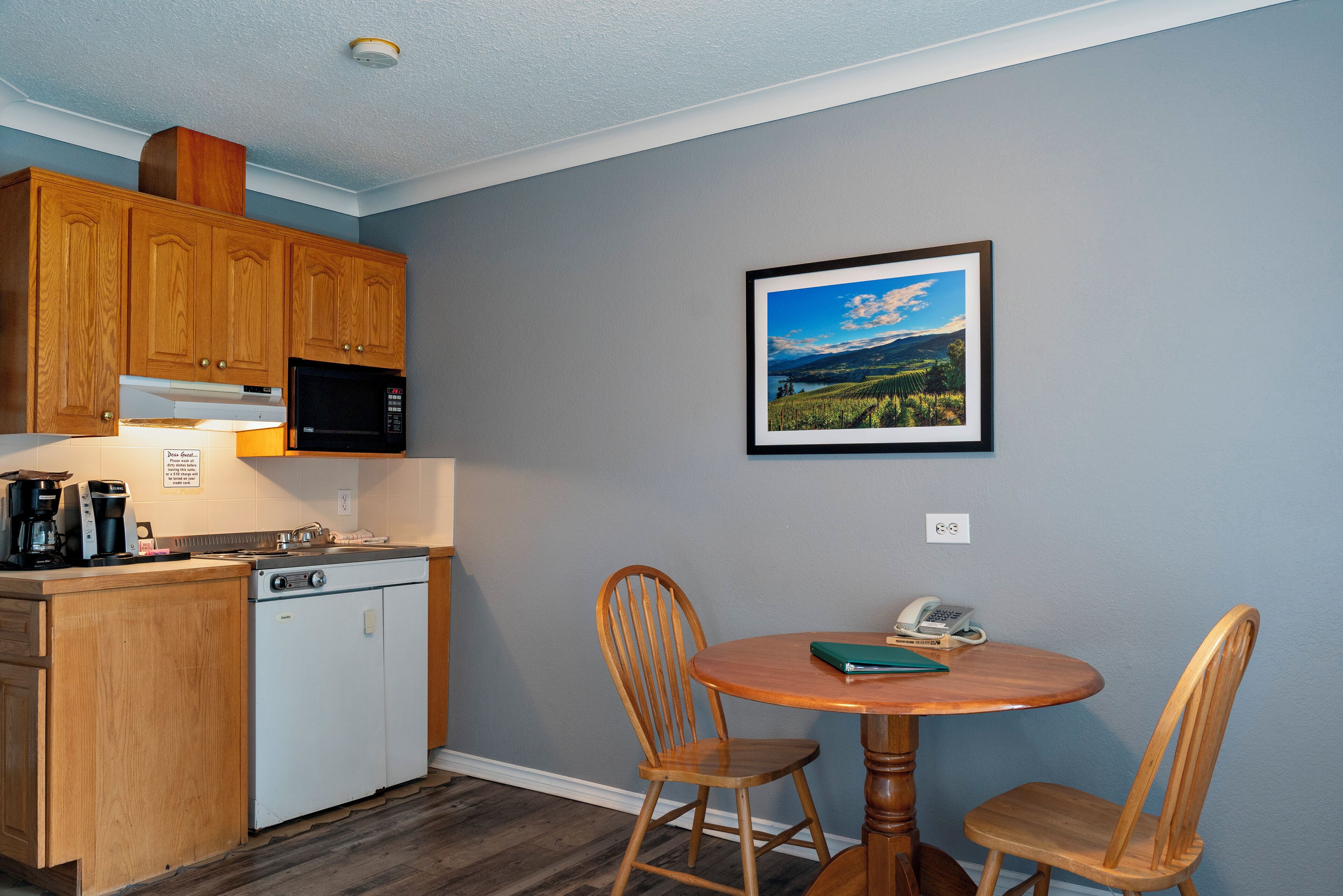 A compact kitchen with wooden cabinets, a white dishwasher, and a black refrigerator. There's a small dining table with two wooden chairs and a framed landscape picture on the wall.
