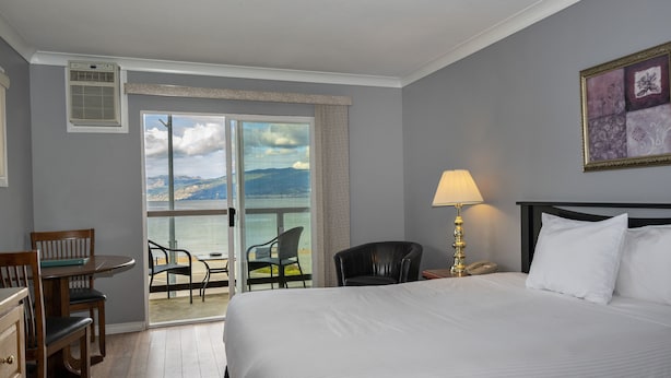 A hotel room with a large bed, a desk with a chair, a lamp, and a view of the sea through a sliding door. - Tiki Shores Inn & Suites (Penticton)