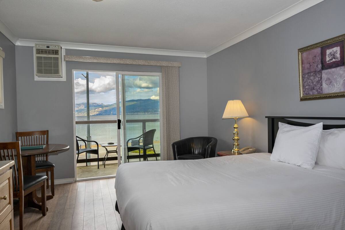 A hotel room with a large bed, a desk with a chair, a lamp, and a view of the sea through a sliding door.