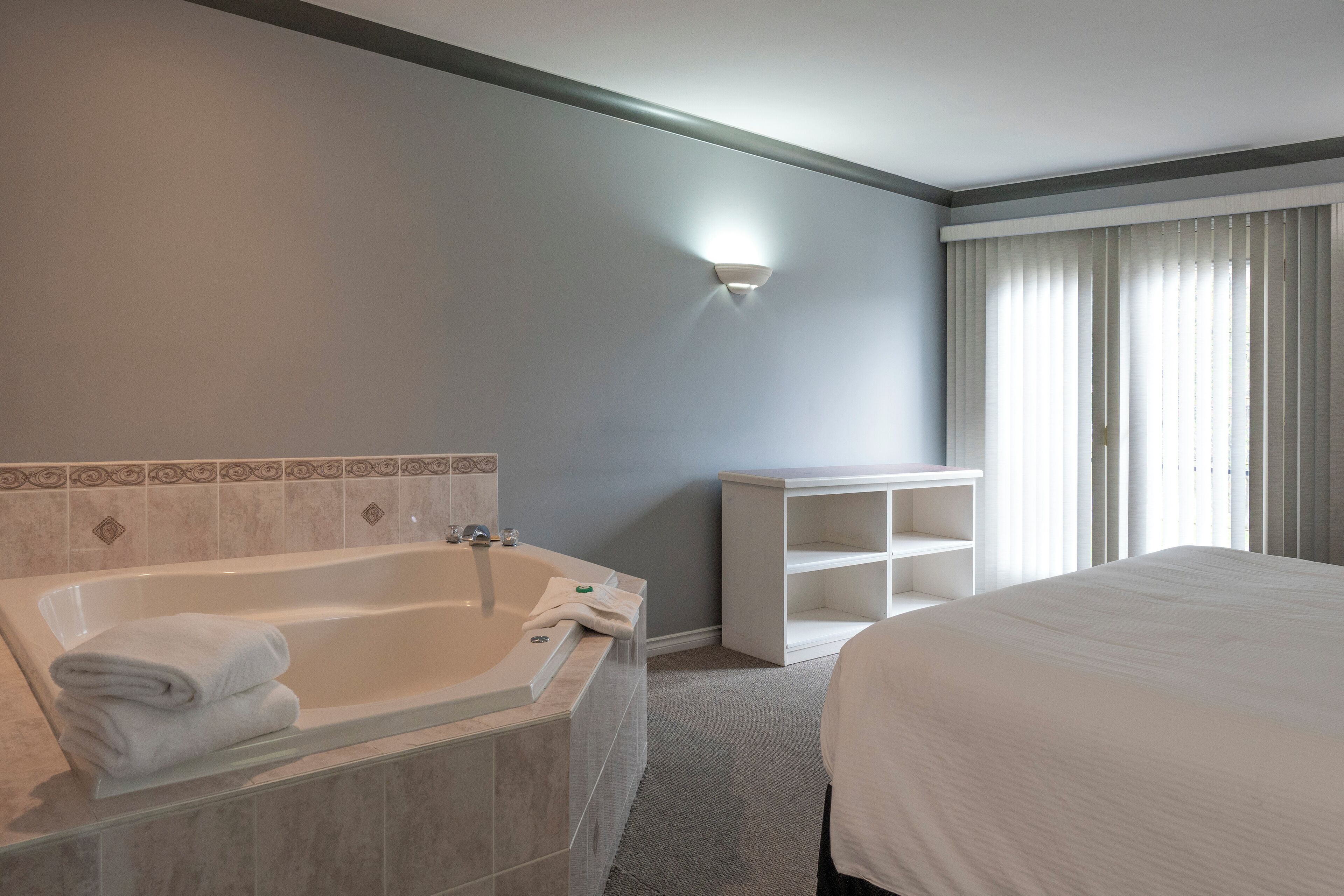 A modern hotel room with a large bed, a bathtub with towels, a small white cabinet, and a window with curtains.